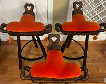 May include: Three antique wooden stools with orange velvet seats. The stools have dark wood frames, decorative carved details, and heart-shaped cutouts at the top. Each stool has a small tassel hanging from the sides. The stools are arranged in a group.