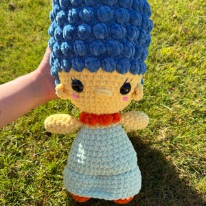 Peut inclure: Une poupée crochetée à la main avec une haute coiffure en laine bleue texturée, un visage jaune pâle avec des yeux de sécurité noirs et des joues roses, un col orange et une robe bleu clair. La figurine est faite de laine épaisse et douce.