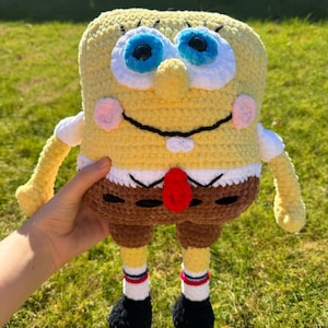 May include: A yellow rectangular crochet plush toy resembling a cartoon character with large blue eyes, a black stitched smile, two white teeth, a red tie, brown shorts, white socks with red and blue stripes, and black shoes.