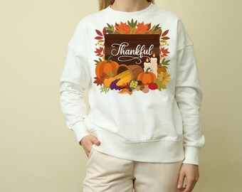 Könnte beinhalten: Weißes Sweatshirt mit einem herbstlichen Design. Die Grafik zeigt Kürbisse, Mais, Trauben und das Wort "Thankful" auf einem Holzschild, umgeben von Herbstblättern. Eine Kerze ist ebenfalls im Design enthalten.