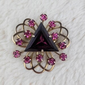 May include: Vintage gold-tone brooch with a large, triangular, dark purple center stone. The brooch is surrounded by smaller, round, pink gemstones and a decorative gold filigree design. A classic piece of jewelry.
