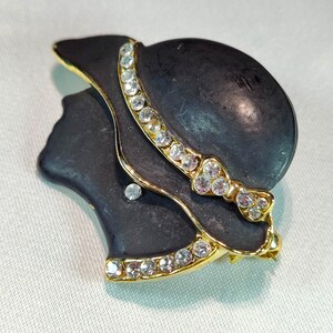 May include: A vintage brooch featuring a black silhouette of a woman wearing a hat. The hat and trim are accented with gold and clear rhinestones. The brooch has a gold-tone border.