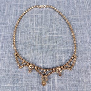 May include: A gold-toned rhinestone necklace with a delicate design. The necklace features a series of small, sparkling rhinestones forming a collar, with larger, square and teardrop-shaped stones at the center. The necklace is displayed on a blue fabric.