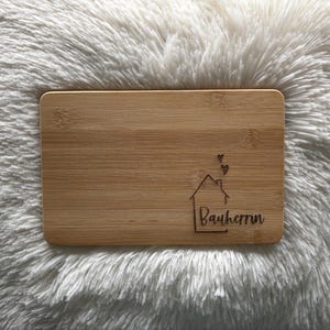 Könnte beinhalten: Ein Holzbrett mit einem Haus- und Herzmotiv und dem eingravierten Text "Bauherrin".