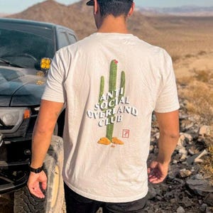 May include: A person wearing a white t-shirt with a green cactus graphic and the text "Anti Social Overland Club". The person is wearing a black baseball cap and black pants. The person is standing in front of a black truck.