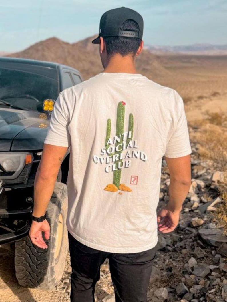 May include: A person wearing a white t-shirt with a green cactus graphic and the text "Anti Social Overland Club". The person is wearing a black baseball cap and black pants. The person is standing in front of a black truck.