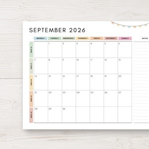 May include: A white paper calendar for September 2026, with a grid layout for daily planning. The calendar includes the days of the week, dates, and space for notes. A pen rests on the right side. The top of the calendar has a decorative banner.