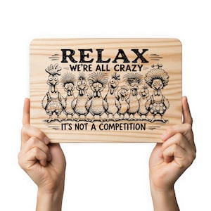 May include: A rectangular wooden sign with the text "RELAX - WE'RE ALL CRAZY - IT'S NOT A COMPETITION" above a cartoon illustration of nine chickens. The chickens have various hairstyles and outfits. The sign is held by two hands.