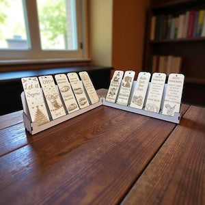 Puede incluir: Dos expositores de madera con marcapáginas grabados. Los marcapáginas presentan varios diseños y textos, incluyendo un árbol de Navidad, un ciervo y coches. El texto incluye frases como "Let it Snow" y "Ride fast, live free." Los expositores están sobre una mesa de madera.