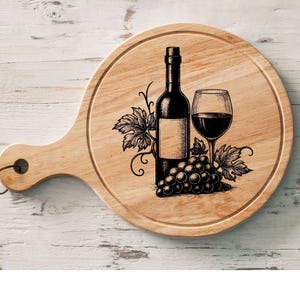 May include: A round wooden cutting board with a handle, featuring a wine bottle, a filled wine glass, and a bunch of grapes. The design is in black and white, with leaves and vines. The board is light brown.