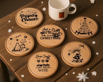 May include: Six round wooden coasters featuring black laser-engraved Christmas designs, including text like Papa Claus, Just a Girl Who Loves Christmas, and Dear Santa Just Bring Wine, alongside a white ceramic mug with a small plaid square graphic.
