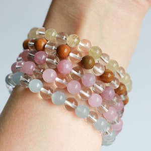 Könnte beinhalten: Eine Sammlung von Armbändern mit Perlen in verschiedenen Farben, darunter Rosa, Hellblau, Klar und Braun. Die Armbänder bestehen aus runden Perlen und sind an einem Handgelenk gestapelt. Die Perlen scheinen aus Edelsteinen und Holz zu bestehen.