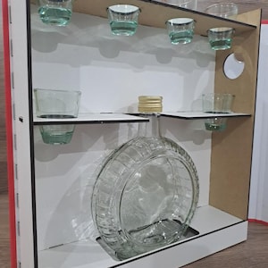 Peut inclure: Un présentoir en bois avec une flasque en verre transparent et des verres à shot. La flasque est ronde avec un motif texturé. Les verres à shot sont remplis d'un liquide vert clair. Le présentoir a un cadre blanc et un dos rouge.