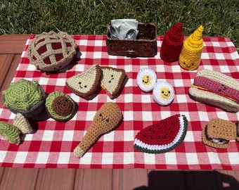 Puede incluir: Una colección de alimentos de juguete tejidos a ganchillo que incluye un pastel, aguacate, sándwich, muslo de pollo, sandía, perrito caliente, huevos duros y botellas de condimentos, todos con caras bordadas sobre un mantel de cuadros rojos y blancos.