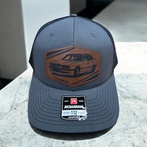 Puede incluir: Una gorra de camionero gris y negra con un parche de cuero marrón que presenta una silueta en blanco y negro de un coche. El parche dice "E30 BMW". La gorra tiene una parte trasera de malla negra y una correa ajustable con una etiqueta blanca que dice "EST. 1970 RICHARDSON ORIGINAL 112 ADJUSTABLE".