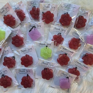 May include: An assortment of small, colorful, air fresheners in various shapes, including clusters of berries and rectangular blocks. Each is individually packaged with a small tag that reads "Thank You" or "Just Because". The fresheners are in shades of red, pink, green, and purple.