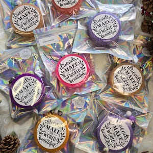 May include: A collection of colorful, round air fresheners in clear, iridescent pouches. Each air freshener has a circular label with the text "friends make the world beautiful" in black script and a decorative leaf design. Various scents are labeled on the pouches.
