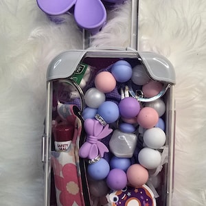 May include: A small, clear plastic suitcase with a purple flower on top. The suitcase is filled with various items, including a purple bow, a donut sticker, and a lip balm. The suitcase is on a white fluffy surface.