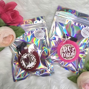May include: Two small, round, pink and red scented sachets with a black string and tag. The pink sachet has the text "Be Mine" in gold glitter lettering. The red sachet has the text "Love" in white glitter lettering. Both sachets are in clear, iridescent bags with a zipper.
