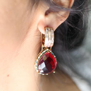 May include: Gold-toned drop earrings with a large, faceted, teardrop-shaped red gemstone. The earrings feature a smaller band of green and clear stones around the red gem and a rectangular band of clear stones at the top.