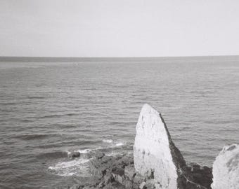 May include: Black and white photograph of a coastal scene. The image captures a view of the ocean meeting a rocky shoreline. A large, light-colored rock formation stands prominently on the right, with the sea and sky filling the background.