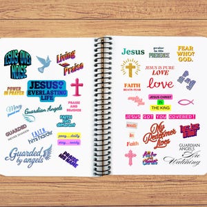 Op de afbeelding: Een open spiraalgebonden notitieboek met diverse kleurrijke stickers met religieuze zinnen zoals "Jezus", "Liefde" en "Geloof". Het notitieboek ligt op een houten oppervlak.