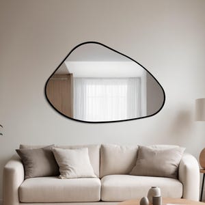 May include: A modern, organically shaped mirror with a black frame is mounted above a cream sofa. The room has a neutral colour scheme, with a round rug, a wooden coffee table, and a potted plant. A lamp and side table are also present.