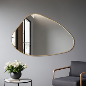 May include: An irregularly shaped gold-framed mirror hangs on a gray wall. Below the mirror is a black armchair with a gray cushion and a small round table with a black frame and a white marble top. A black vase with white flowers sits on the table.