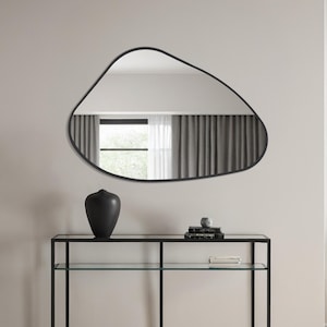 May include: A modern living room scene featuring a black metal and glass console table, a large, irregularly shaped black mirror, and a black vase. The room has neutral-toned walls, a light rug, and a gray armchair.