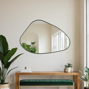 May include: An irregularly shaped mirror with a dark green frame hangs above a wooden bench with a green velvet cushion and gold legs. A large potted plant sits to the left, and a smaller plant is to the right. A neutral-toned rug is on the floor.