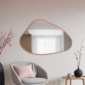 Peut inclure: Miroir mural de forme irrégulière avec un cadre rose pâle. Il reflète une pièce avec une fenêtre et des rideaux blancs. Un fauteuil gris avec un coussin rose est placé en dessous, sur un mur rose clair.