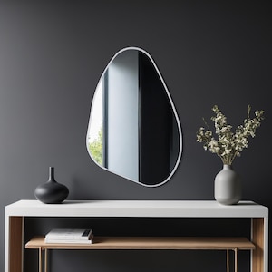 Könnte beinhalten: Ein unregelmäßig geformter Spiegel mit weißem Rahmen hängt an einer dunkelgrauen Wand. Darunter steht ein weißer Konsolentisch mit einer schwarzen Vase, Büchern und einer grauen Vase mit weißen Blumen. Der Spiegel reflektiert ein Fenster.