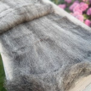 Puede incluir: Primer plano de un material de fibra de lana gris, posiblemente para manualidades o textiles. Las fibras están dispuestas sueltas, creando una apariencia texturizada. El material está colocado sobre una superficie clara, con un fondo borroso de flores rosas.