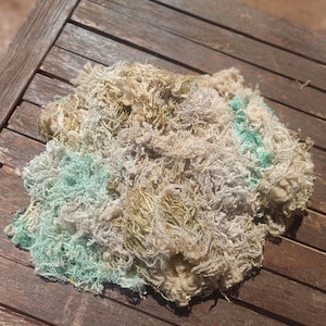 Op de afbeelding: Een hoop getextureerd garen in tinten groen, beige en goud ligt op een verweerd houten oppervlak. Het garen lijkt een mix van vezels te zijn, met een donzige, onregelmatige textuur. De kleuren zijn gedempt en natuurlijk, wat op handwerk duidt.