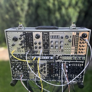 May include: A modular synthesizer system in a silver case with various modules, knobs, and patch cables. The modules have labels like "Braids" and "µPlait". The system is mounted on a black stand, set against a blurred green background.