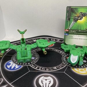 Op de afbeelding: Groene speelgoedfiguren en een kaart met Japanse tekst. De figuren hebben de vorm van mechanische insecten met vleugels en beweegbare delen. De kaart heeft een groen en zwart ontwerp met Japanse tekst en illustraties.