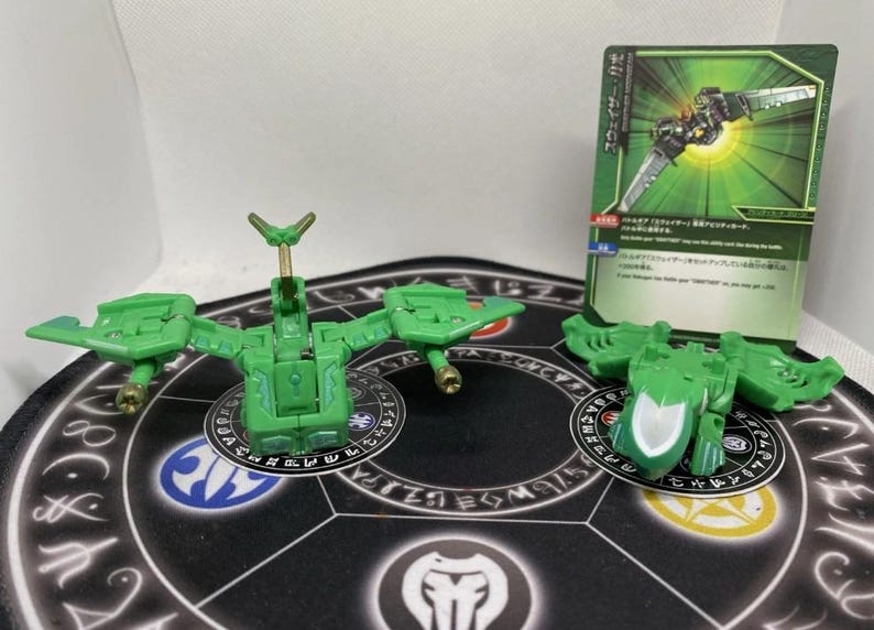 Op de afbeelding: Groene speelgoedfiguren en een kaart met Japanse tekst. De figuren hebben de vorm van mechanische insecten met vleugels en beweegbare delen. De kaart heeft een groen en zwart ontwerp met Japanse tekst en illustraties.