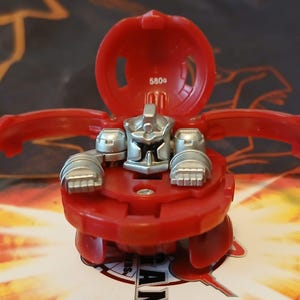 May include: Red and silver toy figure with a hinged top and articulated arms. The figure has a detailed silver face and hands, and the top is marked with "580°". The toy is on a colorful background.