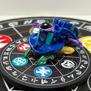 May include: A blue and teal mechanical toy figure with a black dome head and purple accents, featuring articulated limbs and a central panel with a white symbol, resting on a circular black base with white geometric patterns and colored icons.
