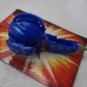 May include: A blue toy ball, partially open, with a segmented design and green and silver accents. The toy is on a card with a red and orange pattern.
