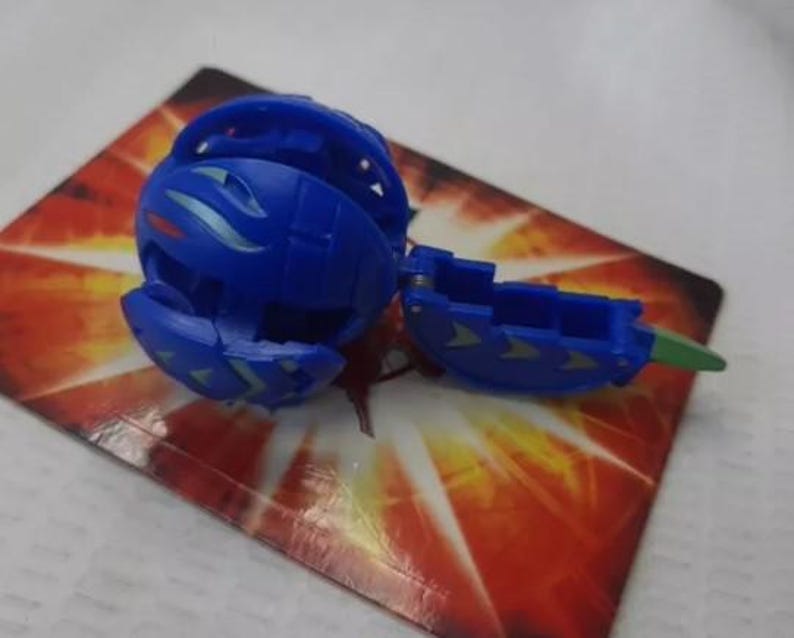May include: A blue toy ball, partially open, with a segmented design and green and silver accents. The toy is on a card with a red and orange pattern.