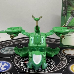 Op de afbeelding: Een groene speelgoedrobot met beweegbare vleugels en gouden accenten. Het staat op een patroonmat, met een groene kaart op de achtergrond. Het ontwerp suggereert een futuristisch thema.