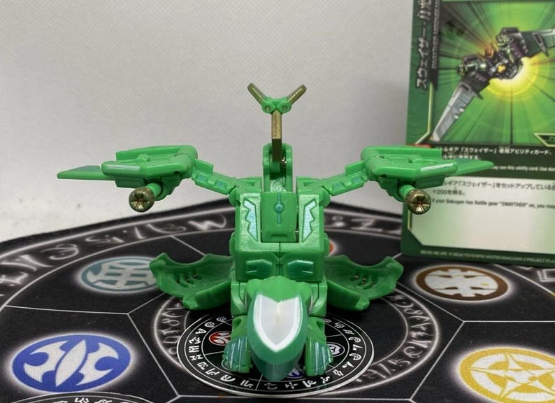 Op de afbeelding: Een groene speelgoedrobot met beweegbare vleugels en gouden accenten. Het staat op een patroonmat, met een groene kaart op de achtergrond. Het ontwerp suggereert een futuristisch thema.