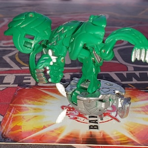Op de afbeelding: Groen en zilveren Bakugan speelgoed met een draakachtig ontwerp. Het speelgoed ligt op een kaart met het woord "Bakugan" erop gedrukt.