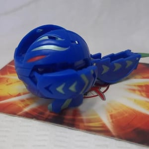 Puede incluir: Una figura de juguete azul, parcialmente abierta, con detalles en verde y plata. El juguete está sobre una tarjeta con un diseño rojo y naranja. El juguete tiene forma de bola con secciones segmentadas y detalles en forma de flecha.