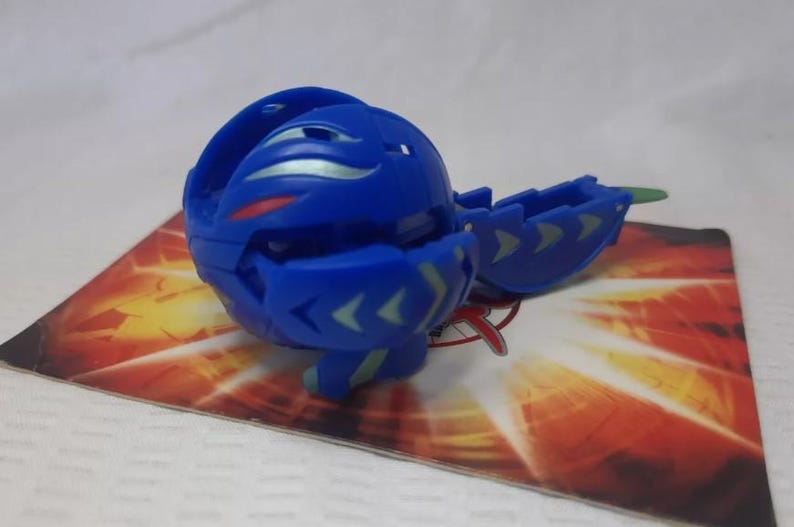 May include: A blue toy figure, partially open, with green and silver accents. The toy is on a card with a red and orange design. The toy has a ball-like shape with segmented sections and arrow-shaped details.