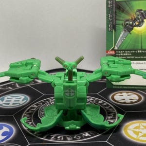 Op de afbeelding: Een groen transformeerbaar speelgoed met beweegbare vleugels en een centraal lichaam, geplaatst op een patroon speelmat. Een ruilkaart met Japanse tekst is zichtbaar op de achtergrond.