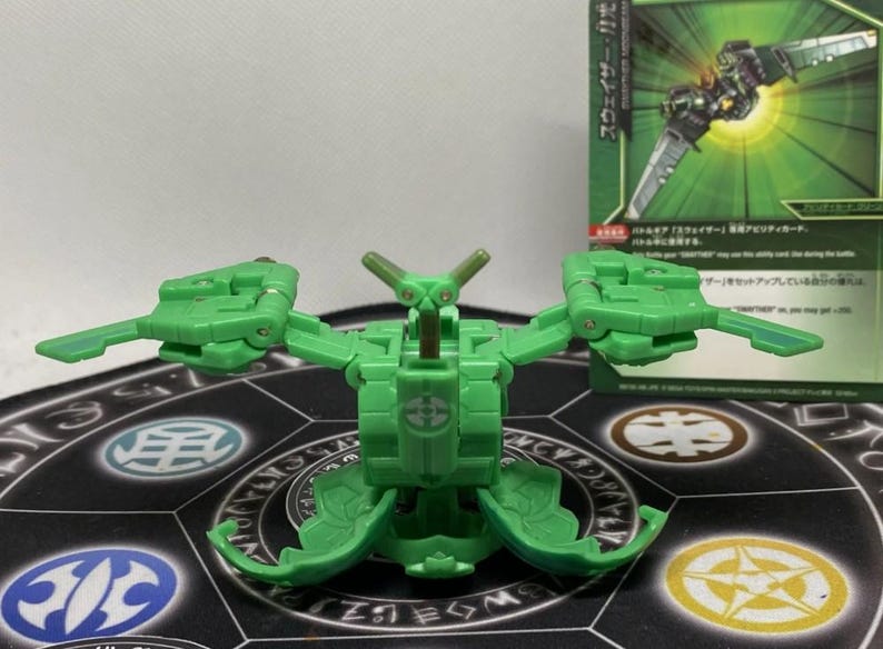 Op de afbeelding: Een groen transformeerbaar speelgoed met beweegbare vleugels en een centraal lichaam, geplaatst op een patroon speelmat. Een ruilkaart met Japanse tekst is zichtbaar op de achtergrond.