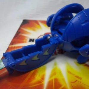 May include: A blue toy figure with a hinged design and green accents. The toy is on a colorful background. The figure has a blade-like extension and is likely part of a collectible toy series.