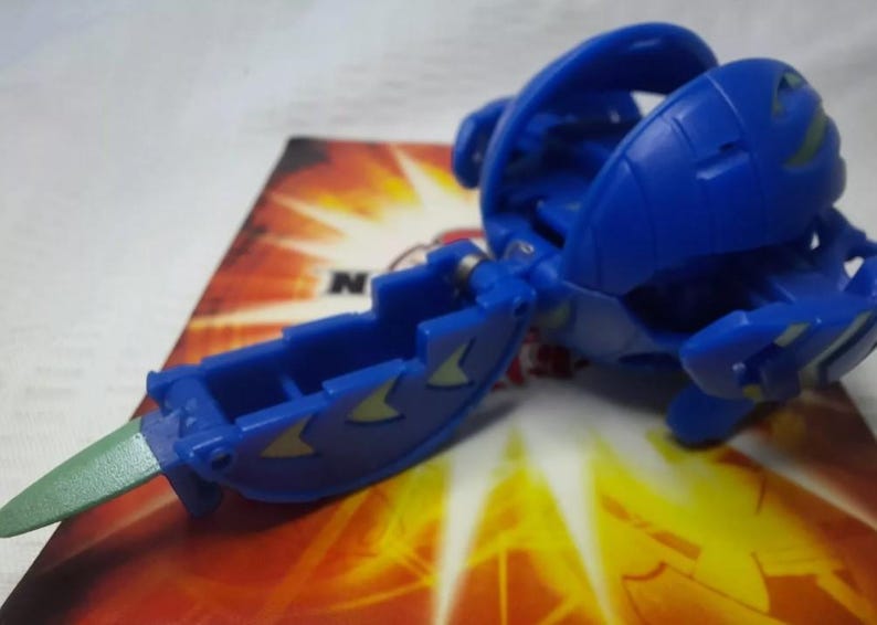 May include: A blue toy figure with a hinged design and green accents. The toy is on a colorful background. The figure has a blade-like extension and is likely part of a collectible toy series.